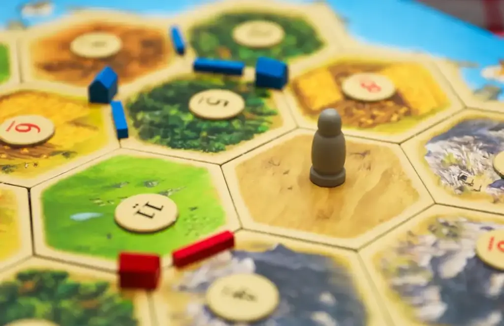 Een Catan-bordspel bestaat uit zeshoekige tegels met verschillende terreinen en nummers. De stukken zijn rood en blauw, met een grijs figuurtje op een woestijntegel.