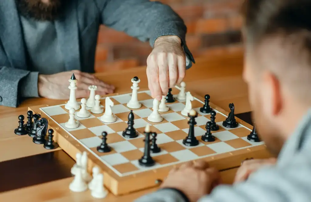 Twee mensen spelen schaak op een houten tafel. De ene persoon verplaatst een witte pion, terwijl de andere intens toekijkt. Geconcentreerde en strategische sfeer.